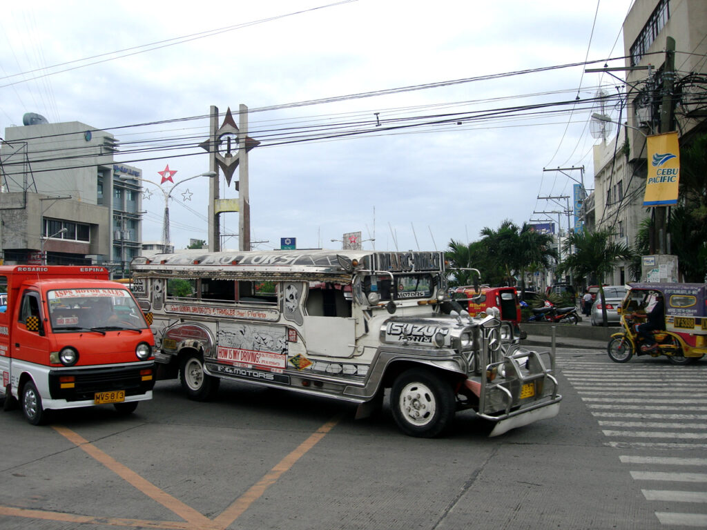 A busy street intersection in General Santos City featuring a chrome-detailed silver and white jeepney with 'ISUZU' visible on its front. The jeepney is parked near a red mini truck or multicab. In the background are urban buildings with utility wires crisscrossing overhead, palm trees lining the street, and a yellow Cebu Pacific banner visible on the right. A city landmark or monument with stars can be seen in the distance, and other vehicles including a tricycle are visible on the road. The scene captures typical Philippine urban transportation.